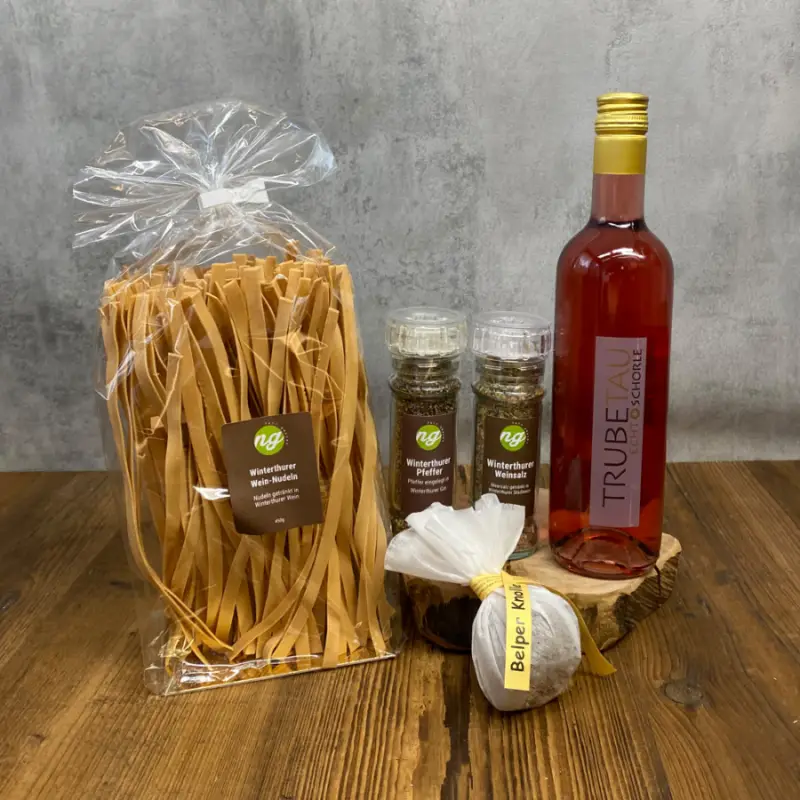 Gift set on a wooden table: a large bag of flat pasta, two pepper mills, a round spice bundle tied with a yellow ribbon, and a tall pink bottle of beverage nearby.