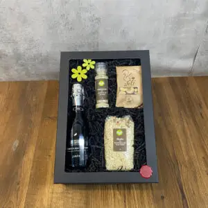 Black windowed gift box showing a bottle of sparkling wine, a spice/pepper grinder, a bag of risotto, crackers, and two yellow flowers on black packing material.