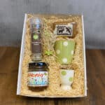 Gift box with straw filling containing a spice grinder labeled 'Gewürzmischung für das Ei', a jar of honey, a green polka-dot mug, a white mug with green dots, and a small decorative soap, plus dried plant accents for a gift set.