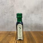 Small dark glass bottle with a green cap on a wooden table; label suggests balsamic vinegar, reading 'Selezione buonsapore' and 'Aceto balsamico'.