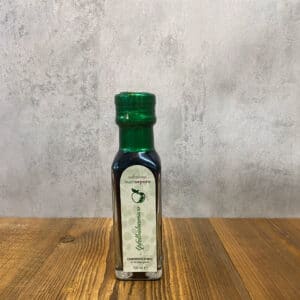 Small dark glass bottle with a green cap on a wooden table; label suggests balsamic vinegar, reading 'Selezione buonsapore' and 'Aceto balsamico'.