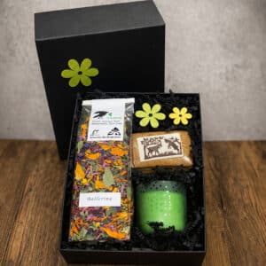 Black gift box opened to reveal a colorful dried herb tea labeled 'Ballerina', a deer-illustrated soap, and a green dotted candle, with yellow felt flowers as decoration