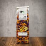 Tall transparent bag of dried edible flower petals with a white label and green logo, labeled 'Fleurs de salade', on a wooden table against a gray wall.