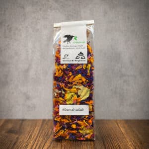 Tall transparent bag of dried edible flower petals with a white label and green logo, labeled 'Fleurs de salade', on a wooden table against a gray wall.