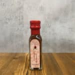 Small glass bottle of pomegranate balsamic vinegar with a red cap on a wooden table surface, pink label visible