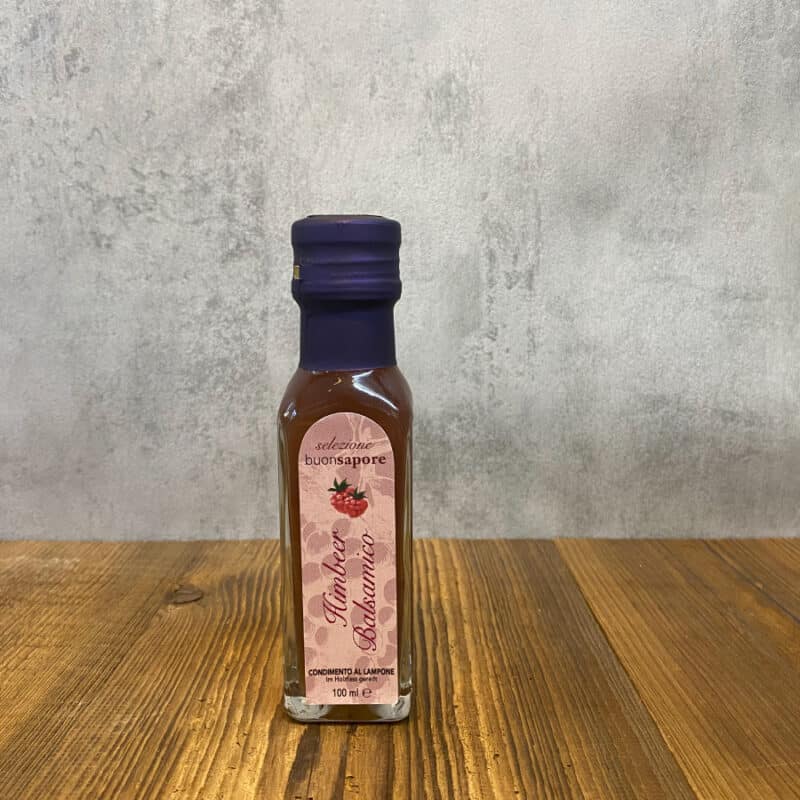 Small glass bottle with a dark purple cap on a wooden table; pink label shows a berry illustration and reads 'Selezione buonsapore' with '100 ml' at the bottom, suggesting a flavored sauce bottle.