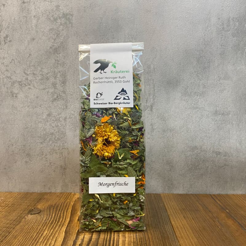 Tall clear bag of Morgenfrische herbal tea with dried greens and yellow flowers on a wooden surface; label shows Kräuterei branding and Swiss bio logos.