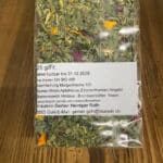 Transparent bag of dried herbal tea with a white label listing ingredients and producer information in German on a wooden surface. Includes notes like 'Kräuterei Gerber Heiniger Ruth' and contact details.