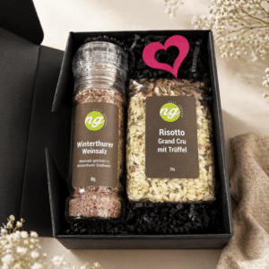 Gift box containing two labeled food products: Winterthurer salt grinder and Risotto Grand Cru mit Trüffel, with a pink heart ornament on black filler material inside the box