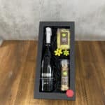 Gift box containing a bottle of prosecco, risotto mix, and seasoning jars on a wooden table, with yellow flower decorations and a red wax seal at the corner.
