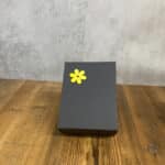 Dark gray gift box with a bright yellow flower sticker on the lid, resting on a wooden surface.