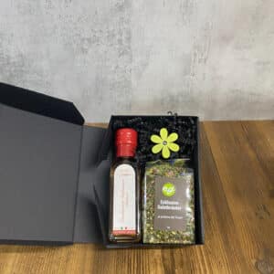 Gift box with a red-capped bottle and a bag of salad herbs, surrounded by black shreds and a bright yellow decorative flower.