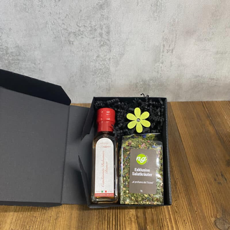 Gift box with a red-capped bottle and a bag of salad herbs, surrounded by black shreds and a bright yellow decorative flower.