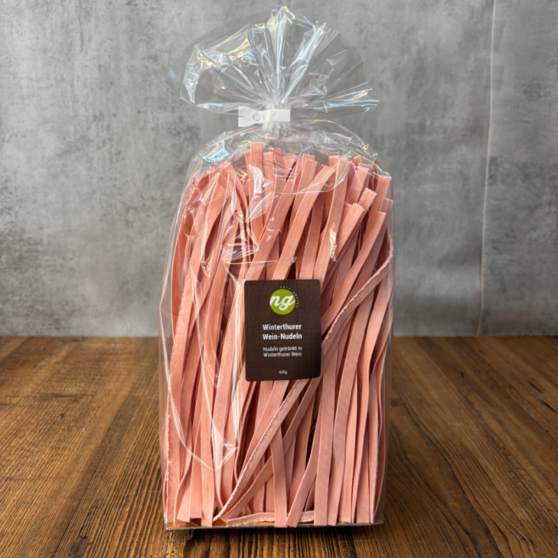 Bag of pink flat pasta ribbons labeled Winterthurer Wein-Nudeln inside a clear gift bag on a wooden table, ready for sale or gift.